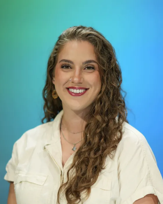 A woman with curly hair smiles while wearing a white shirt against a blue and green background.
