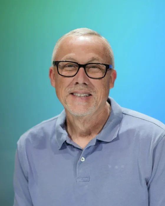 A man with glasses smiles while wearing a light purple polo shirt against a gradient blue background.