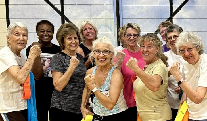 A group of women poses with resistance bands, smiling and flexing their arms in a fitness setting.