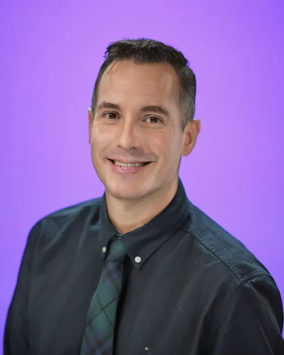 A man smiles while wearing a dark shirt and a patterned tie, with a purple background.