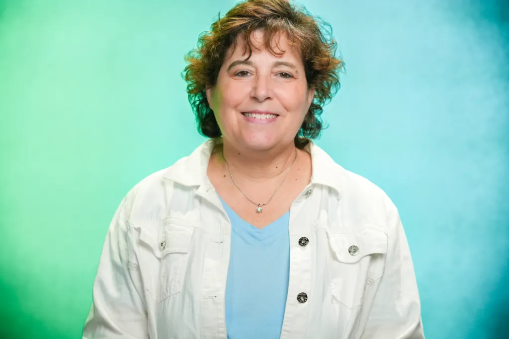 A woman with curly hair smiles while wearing a white jacket over a blue shirt, set against a green background.