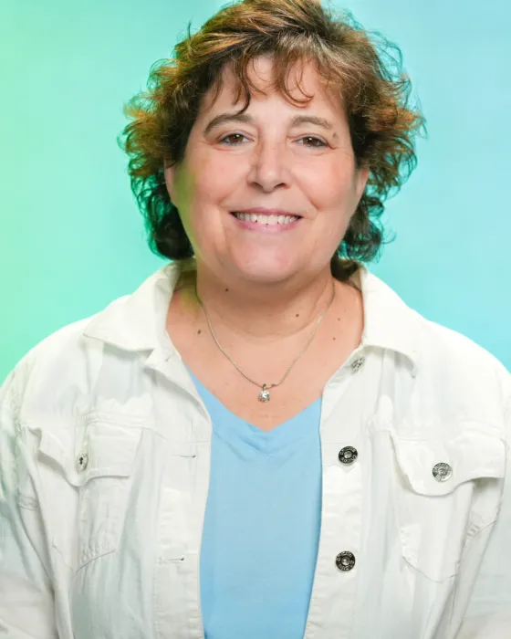 A woman with curly hair smiles while wearing a white jacket over a blue shirt, set against a green background.