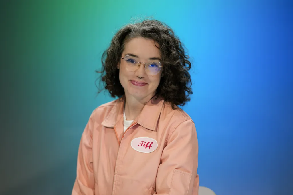 A person with curly hair and glasses smiles while wearing a pink outfit with a name tag that reads "Tiff.