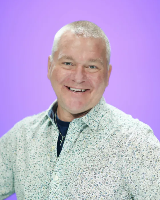 A man with a short haircut smiles while wearing a patterned shirt against a purple background.