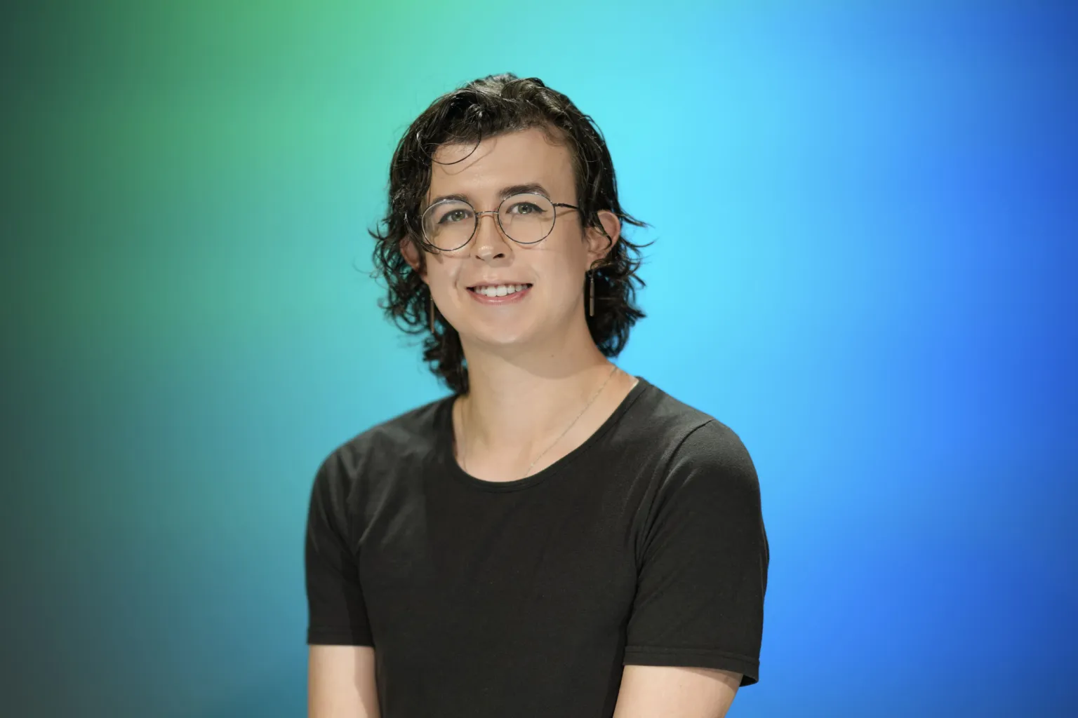 A person with curly hair and glasses smiles while wearing a black shirt, set against a blue-green background.