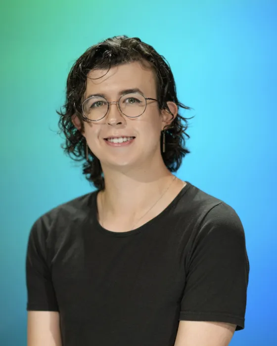 A person with curly hair and glasses smiles while wearing a black shirt, set against a blue-green background.