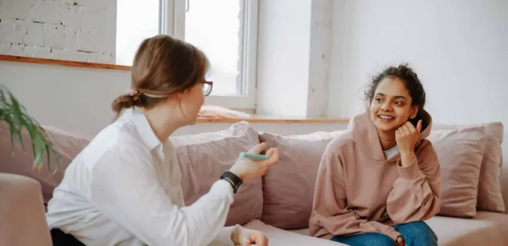 Two women are sitting on a couch, engaged in conversation, with one speaking and the other listening and smiling.