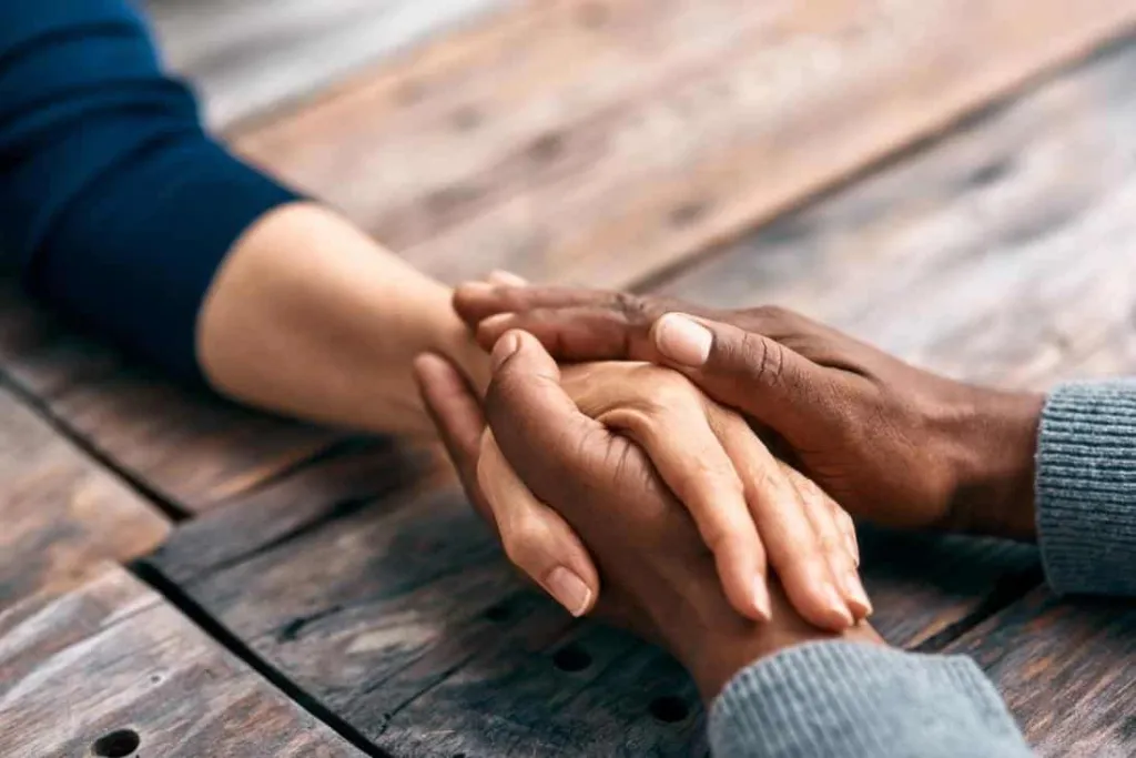 Two hands are clasped together on a wooden surface, one hand resting on top of the other.
