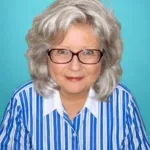 A woman with gray hair and glasses smiles while wearing a blue and white striped shirt against a turquoise background.