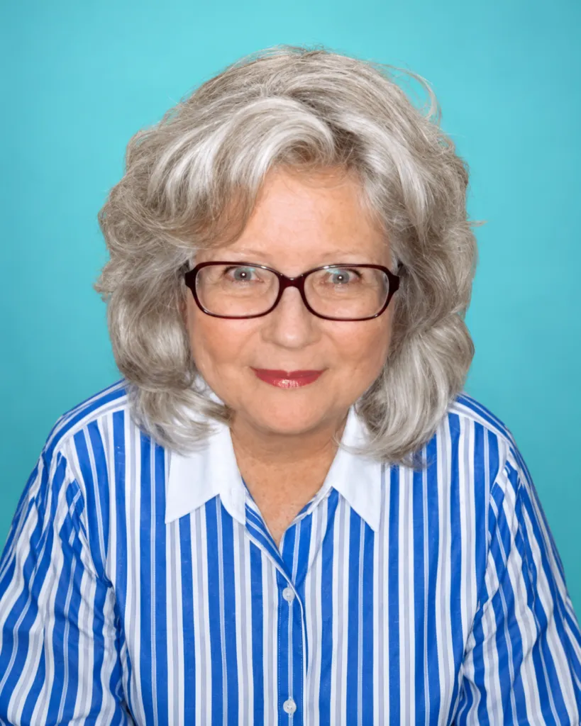 A woman with gray hair and glasses smiles while wearing a blue and white striped shirt against a turquoise background.