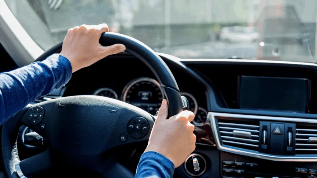 Hands grip the steering wheel of a car while driving, with a dashboard and road visible in the background.