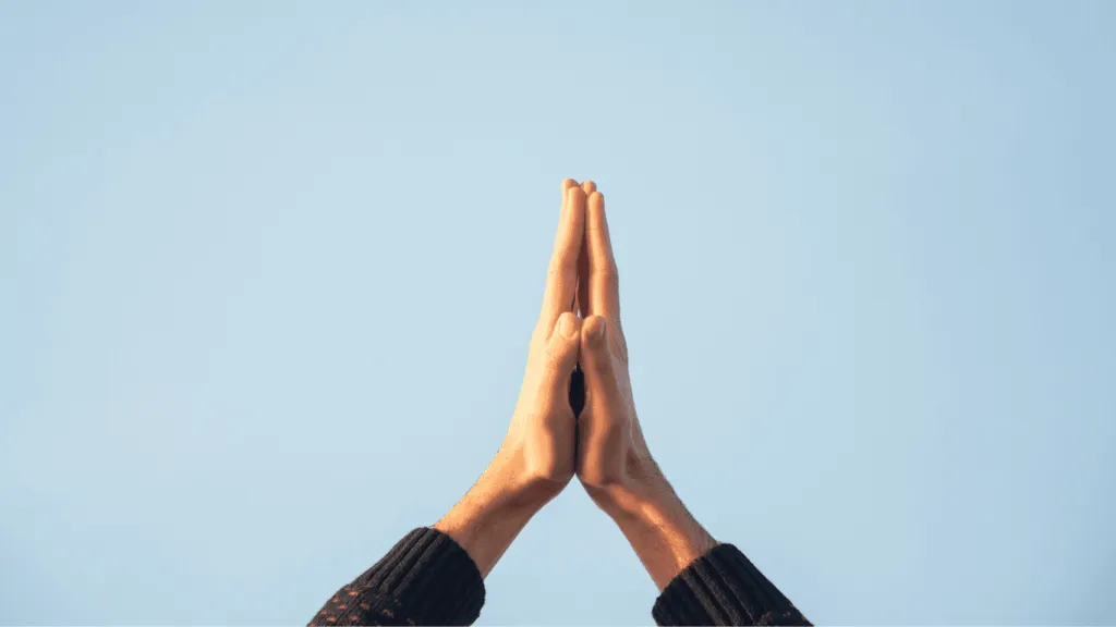 Hands are clasped together in prayer against a clear blue sky.