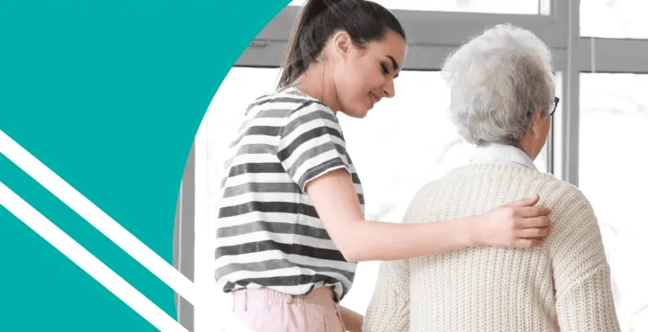 A young woman assists an older woman while walking, both facing away from the camera in a bright indoor setting.