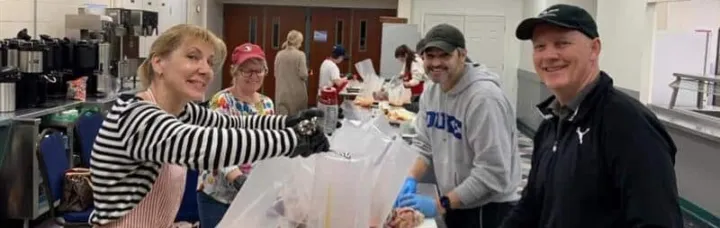 Volunteers prepare food together in a kitchen, smiling and working at a long table with various food items.