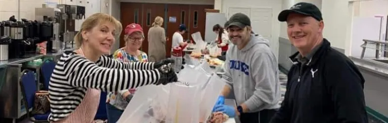 Volunteers prepare food together in a kitchen, smiling and working at a long table with various food items.