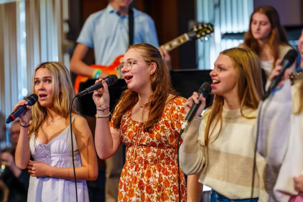 Youth singers perform with microphones on stage, accompanied by a guitarist in the background.