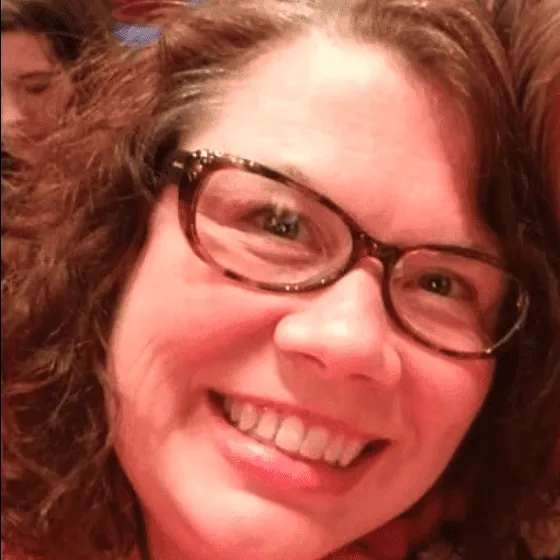 A woman with curly hair and glasses smiles at the camera, with a blurred background of other people.