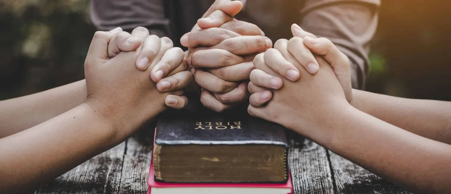 Hands are clasped together in prayer over a closed Bible on a wooden table.