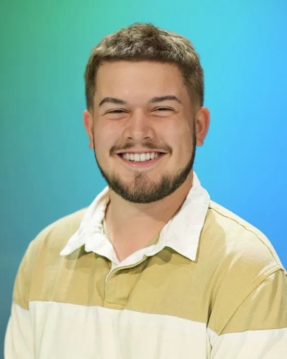 A young man with a beard smiles while wearing a striped shirt against a blue-green background.