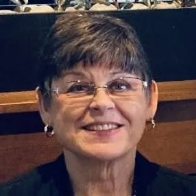 A woman with short hair and glasses smiles while sitting indoors, with a wooden surface and decorative items in the background.
