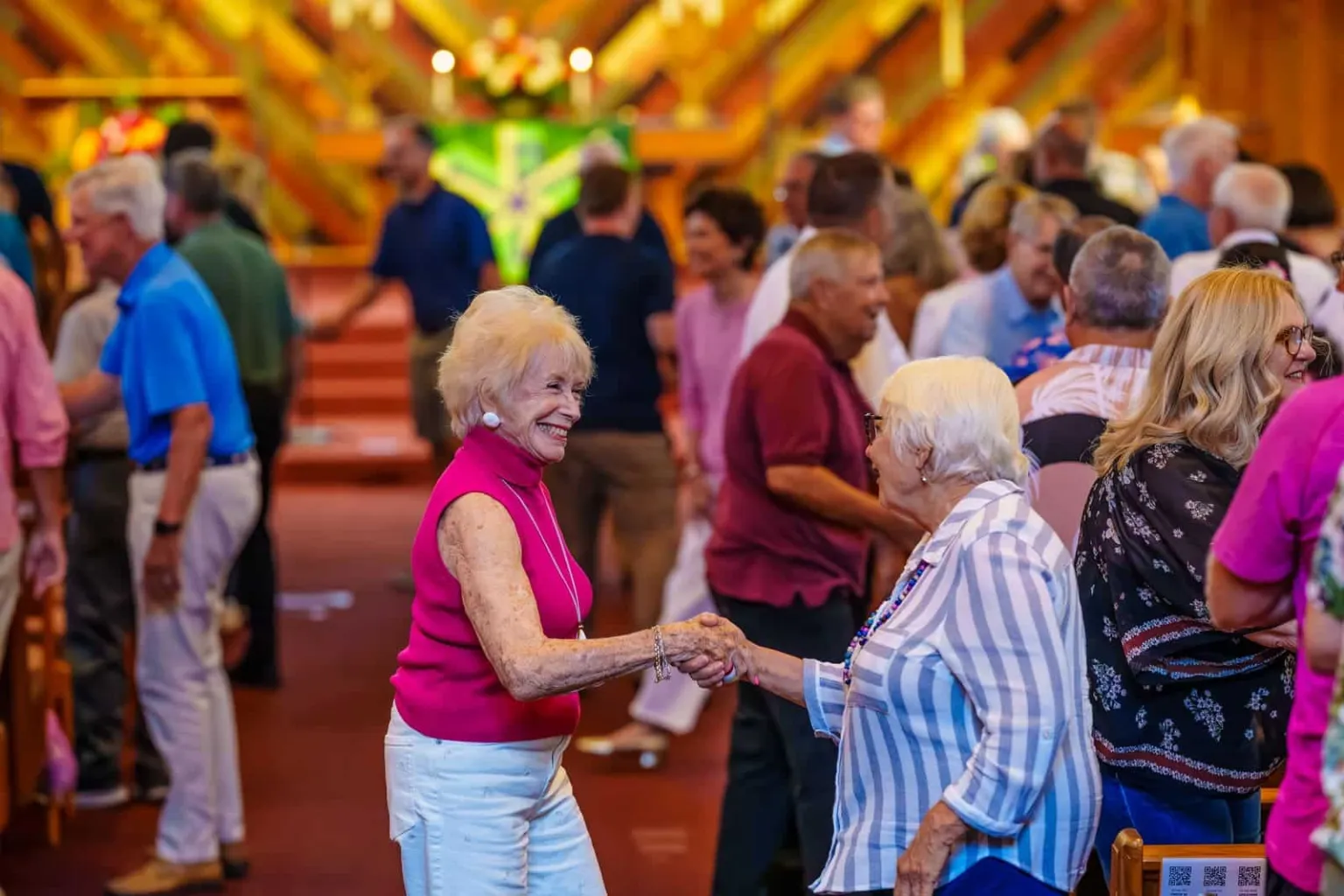 Two women shake hands and smile while interacting with a crowd of people in a church setting.