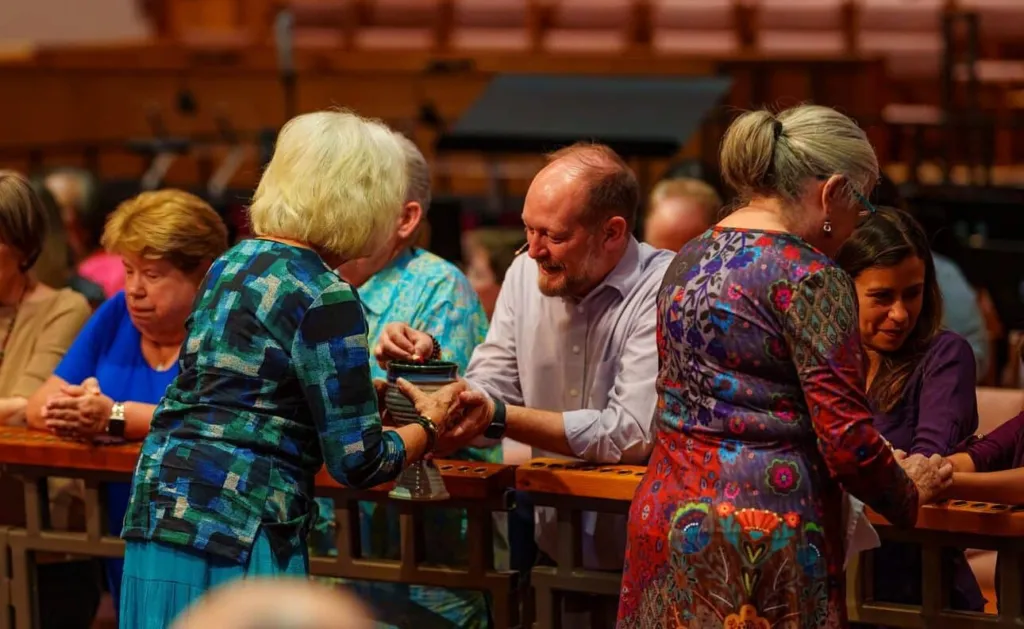 People are engaged in conversation and exchanging items at a long table in a church setting.
