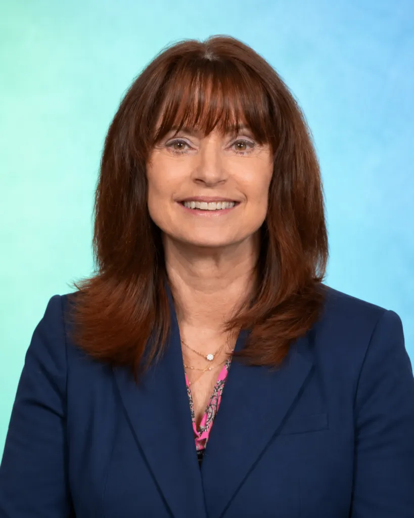 A woman with long brown hair smiles while wearing a blue blazer over a floral top, against a light blue background.