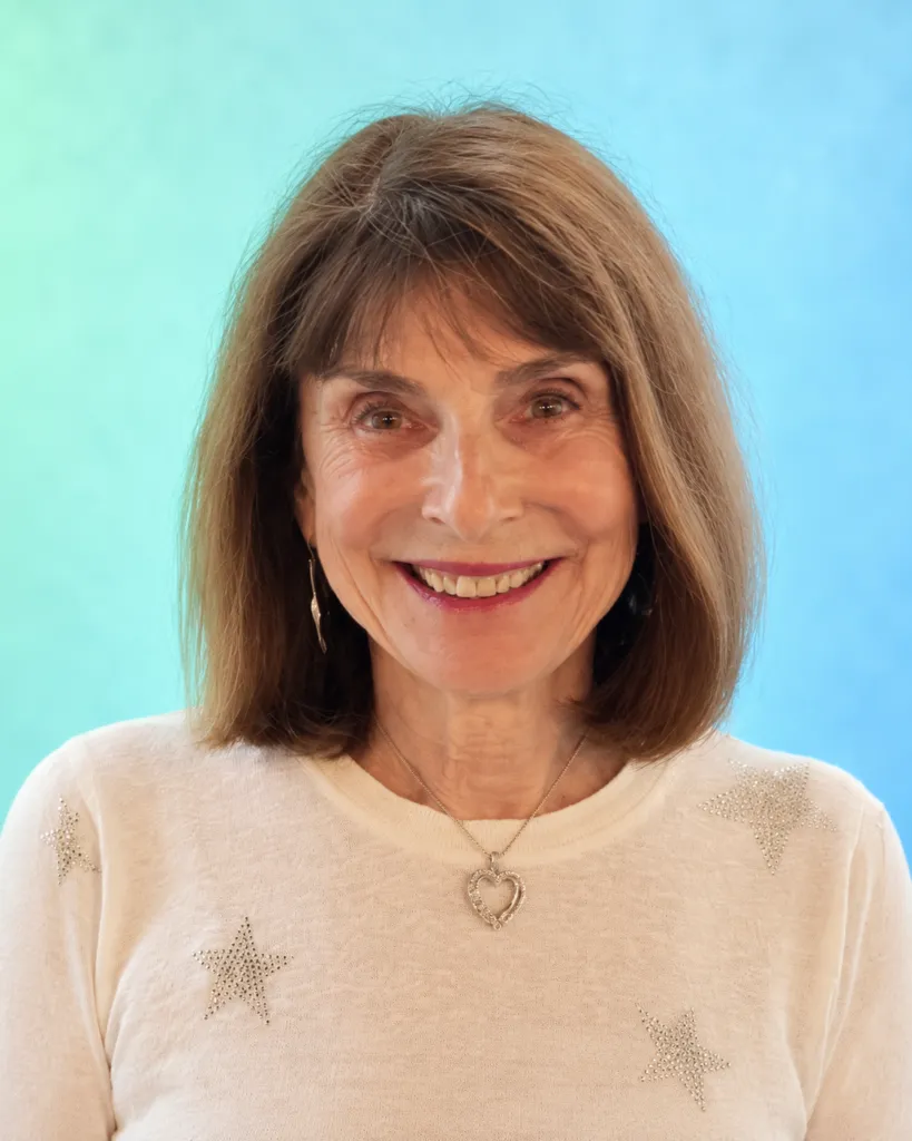 A woman smiles while wearing a star-embellished sweater and a heart necklace against a blue background.