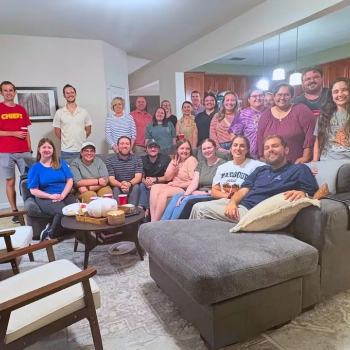 A large group of young adults gathers in a living room, smiling and posing for a photo.