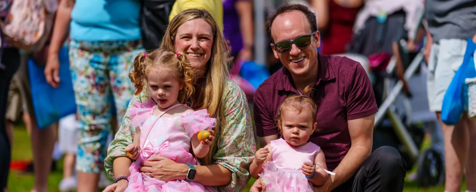 A family poses for a photo outdoors, with two young girls in pink dresses sitting on their parents' laps.