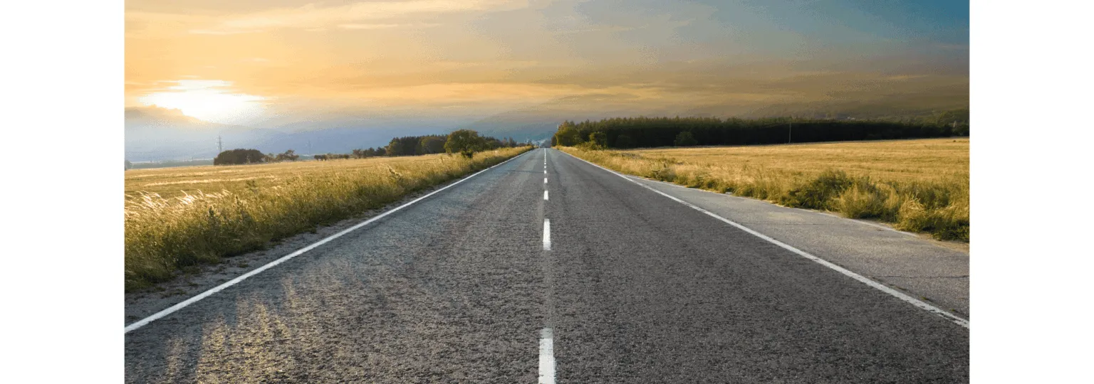 A long, straight road stretches into the horizon, bordered by fields and a sunset sky.