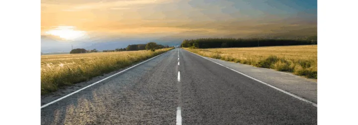 A long, straight road stretches into the horizon, bordered by fields and a sunset sky.