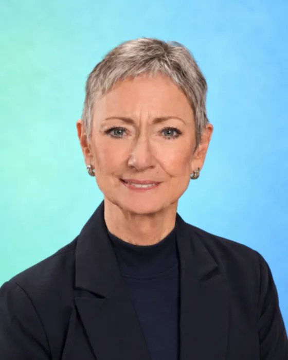 A woman with short hair is smiling while wearing a black blazer against a blue-green background.