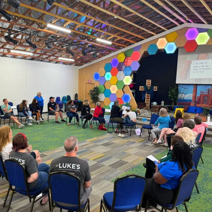 Participants are seated in a circle, engaging in a discussion in a colorful room with a decorative wall.