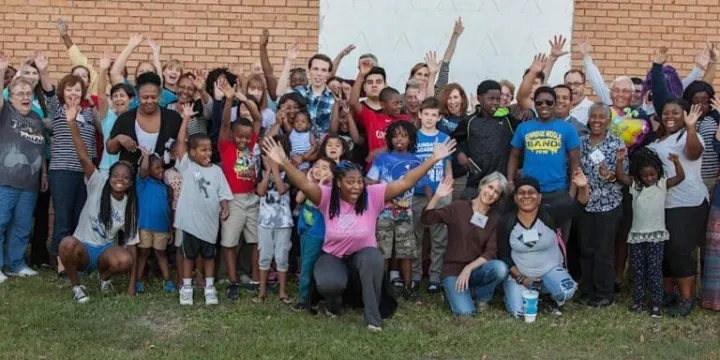 A large group of people of various ages poses outdoors, smiling and waving with their hands raised.