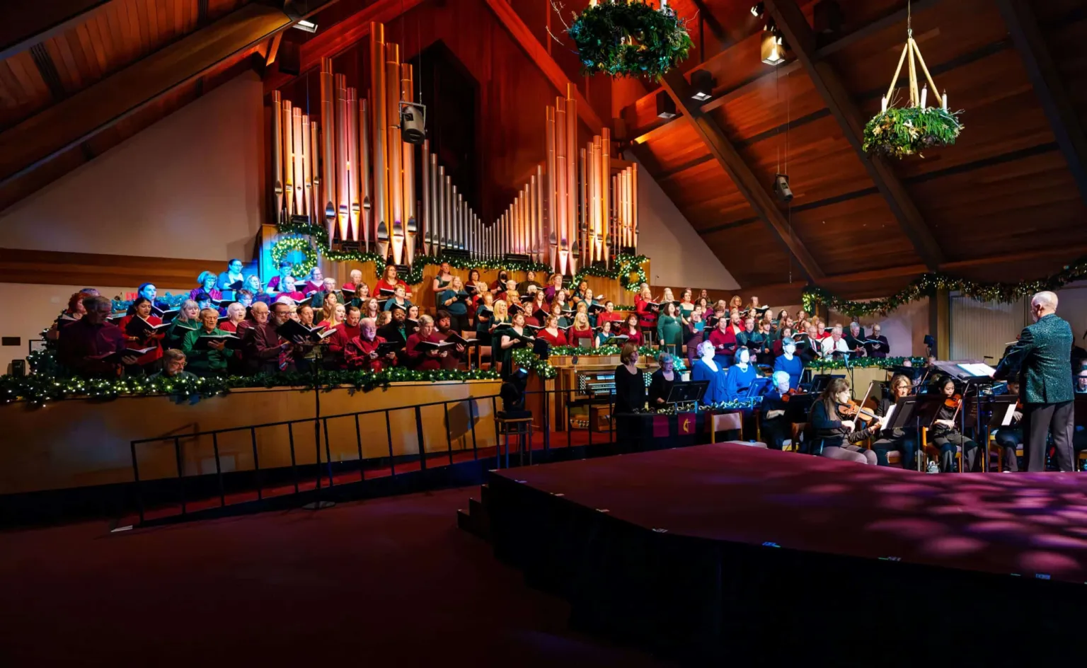 A choir performs on stage with musicians, surrounded by festive decorations and a large organ in the background.