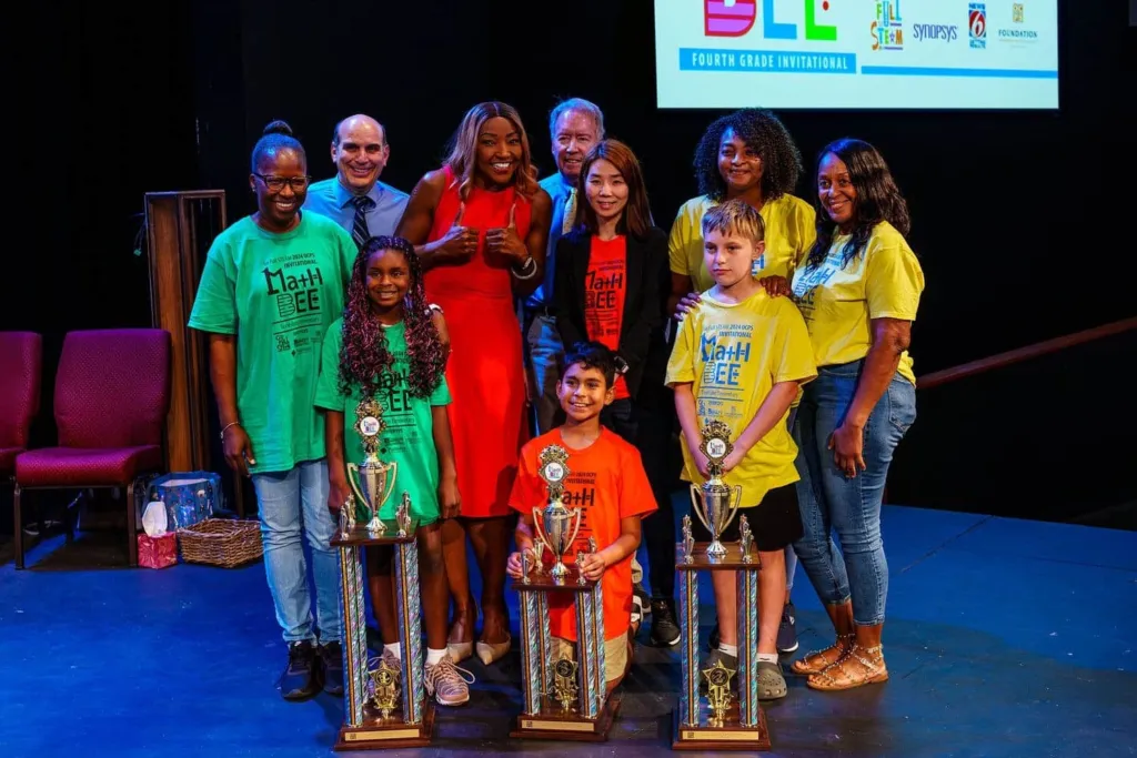 Participants celebrate with trophies after a Math Bee competition, posing together on stage.