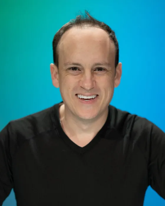 A man in a black shirt smiles while leaning forward against a blue-green background.