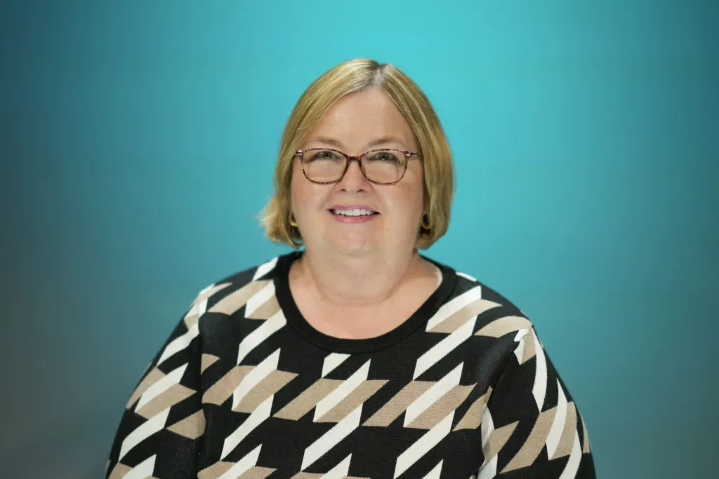 A woman with short blonde hair and glasses smiles while wearing a patterned black and beige sweater against a blue background.