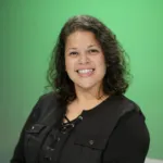A woman with curly hair smiles while seated in front of a green background.
