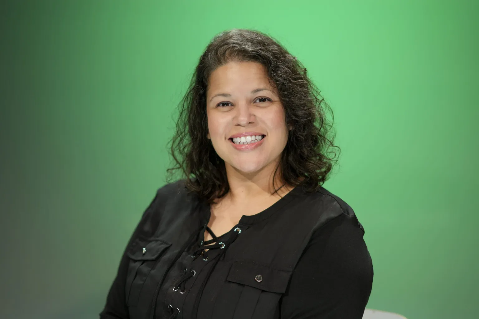 A woman with curly hair smiles while seated in front of a green background.