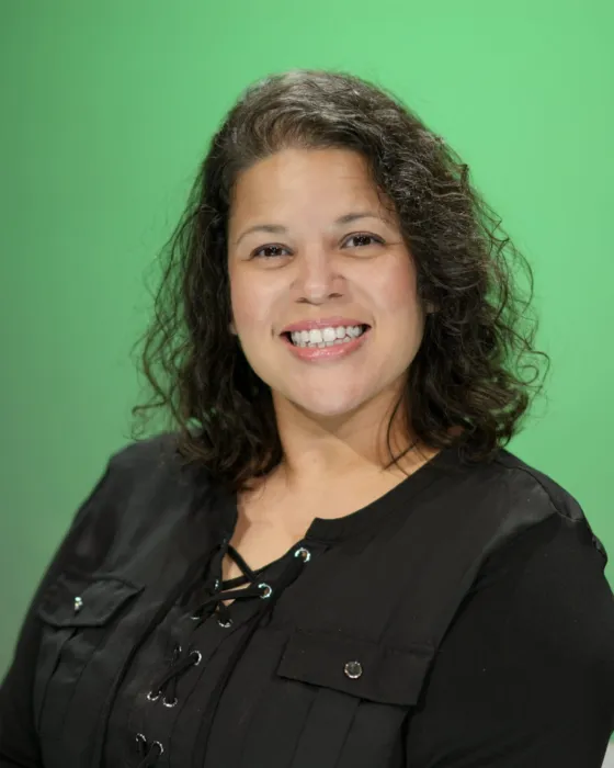 A woman with curly hair smiles while seated in front of a green background.