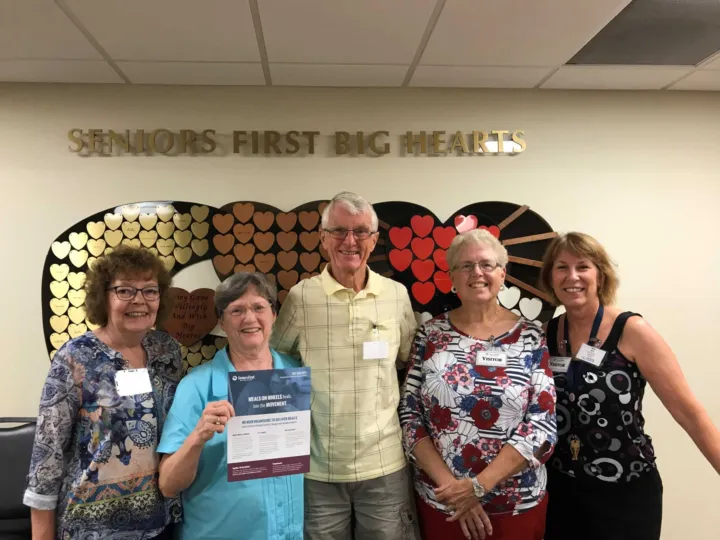 Five people stand together, holding a Meals on Wheels flyer, in front of a wall decorated with hearts.
