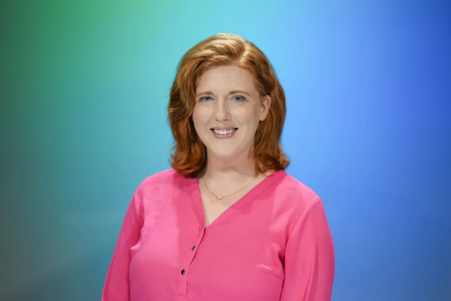A woman with red hair smiles while wearing a pink blouse against a blue and green background.