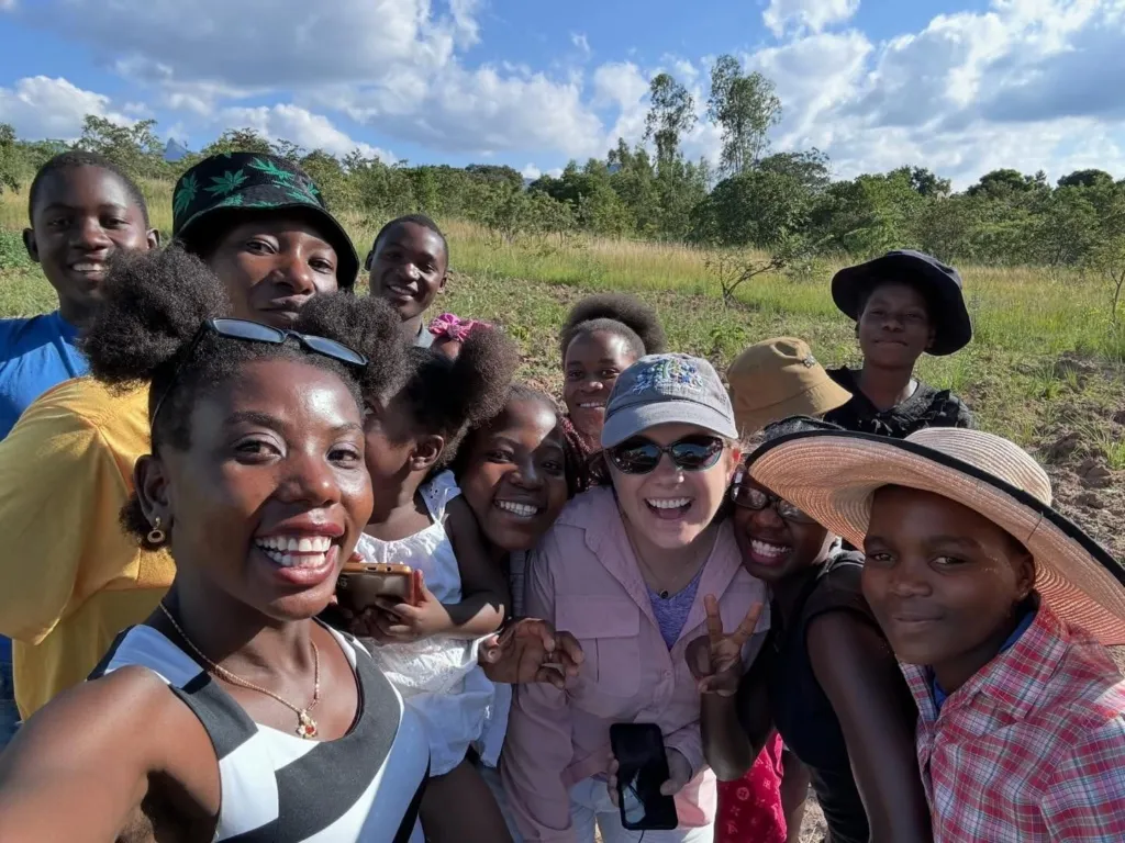 A group of young people and an adult pose for a selfie outdoors, smiling and enjoying their time together.