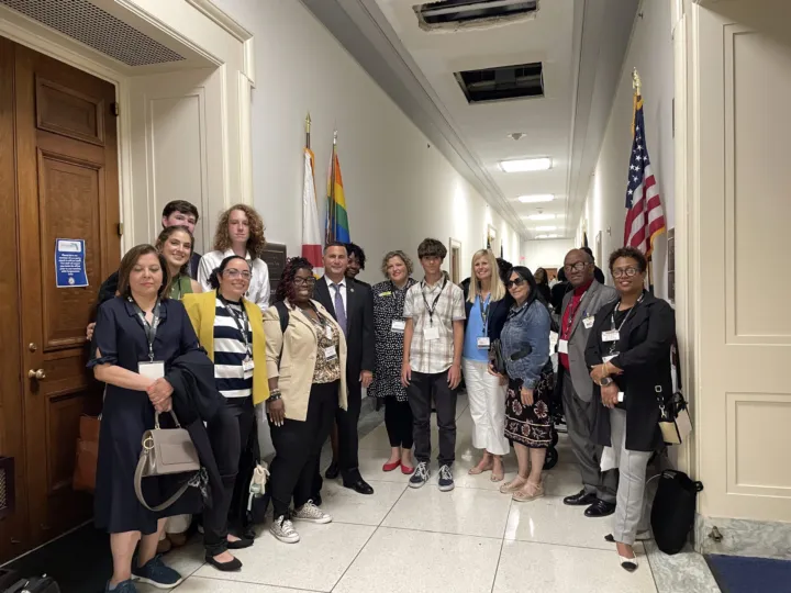 A diverse group of individuals poses together in a hallway, with flags and doors visible in the background.