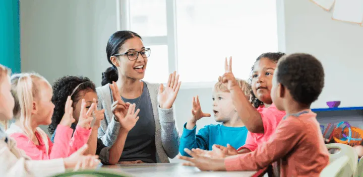 A teacher engages with a group of children at a table, all raising their hands and participating in an activity.