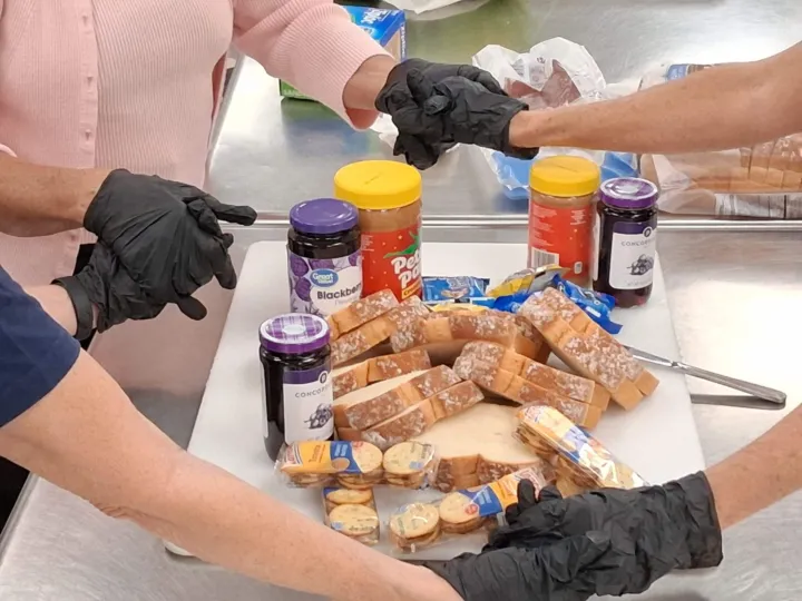 Hands in gloves are preparing sandwiches with bread, peanut butter, and jelly on a table.
