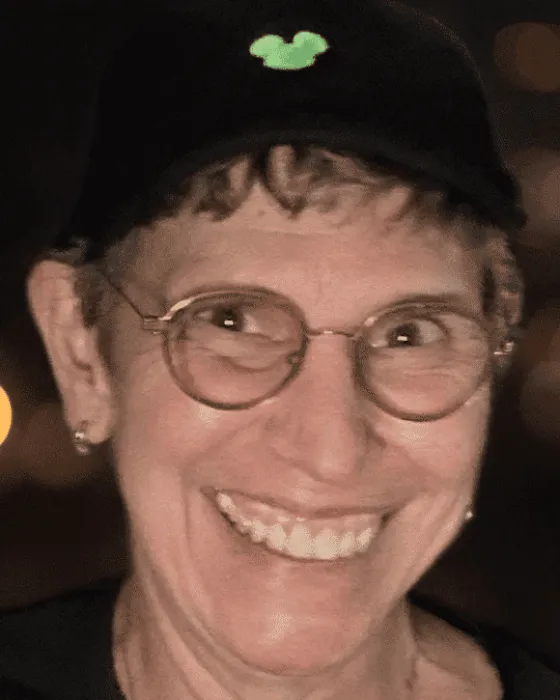 A smiling person wearing glasses and a black cap with a green Mickey Mouse emblem stands against a blurred background.