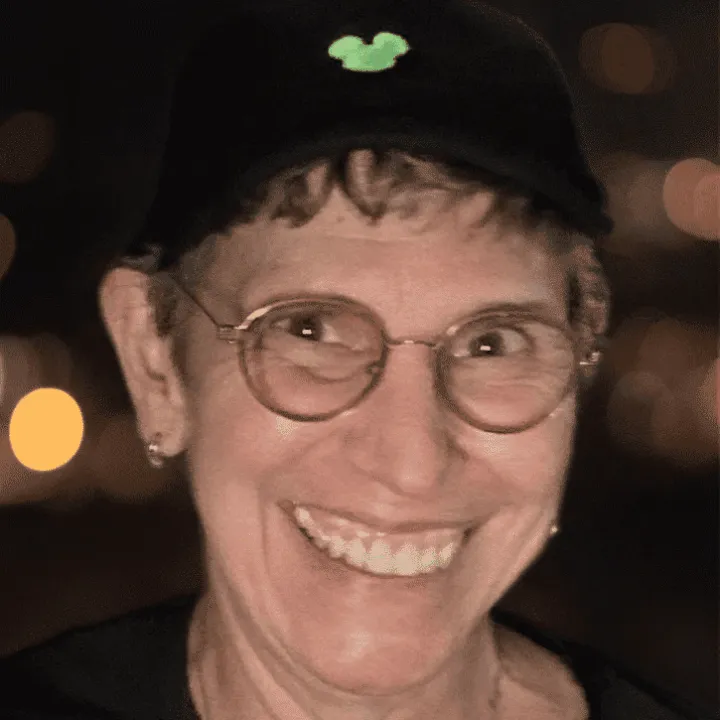 A smiling person wearing glasses and a black cap with a green Mickey Mouse emblem stands against a blurred background.